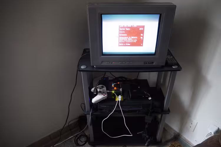 In a bedroom corner is a black three-tier display rack with cables hanging messily around and with a gray CRT television on the top shelf that is turned on and displaying the MiSTer FPGA menu, a black VCR on the middle shelf with a MiSTer FPGA unit, peripherals, and white game controller, and a black PlayStation controller on the bottom shelf